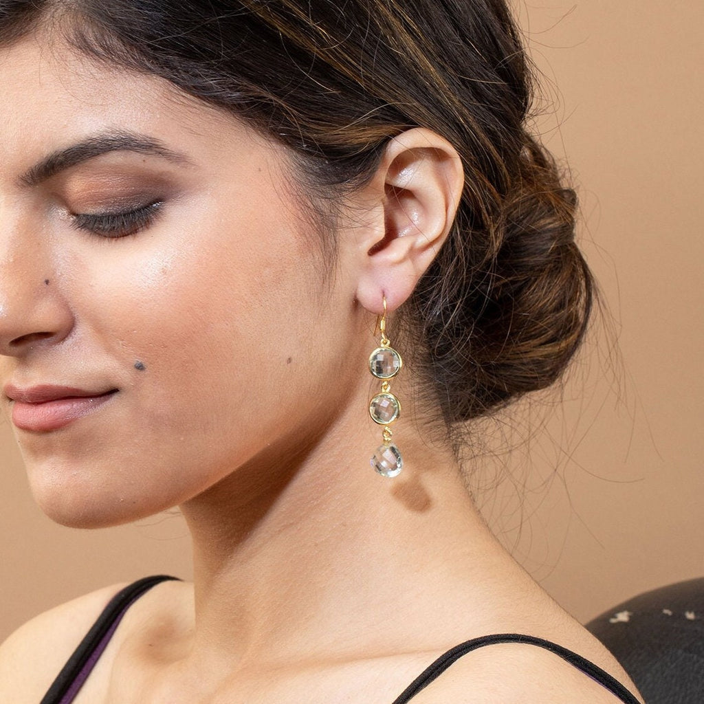May include: Gold-toned drop earrings featuring three clear, round gemstones. The gemstones are suspended from a delicate gold hook, creating a cascading effect. The earrings are shown on a person's ear, highlighting their elegant design.